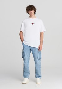 Biała koszulka z krótkim rękawem z grafiką czerwonej róży i czarnym napisem. Jasnoniebieskie spodnie cargo z wieloma kieszeniami. Beżowe sneakersy.