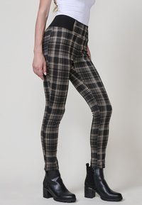 Slim-fit hlače v črno-bež karirastem vzorcu, opremljene s črnim elastičnim pasom. V kompletu s črnimi gležnjarji na peto z okrasnimi studenčki.