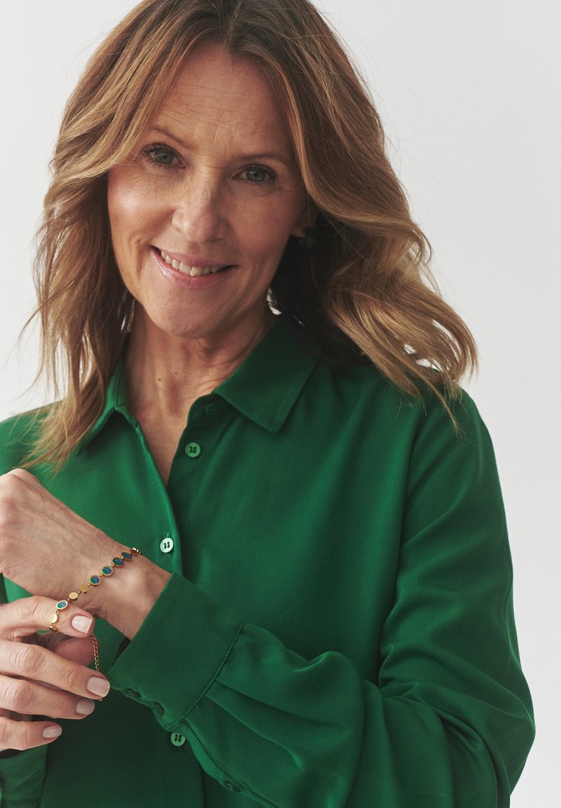 Bracciale d'oro con gemme ovali multicolori, indossato su un polso, abbinato a una camicia verde con colletto e dettagli a bottoni.