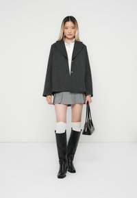 Veste en laine grise avec un large col, fermeture à boutons, associée à une jupe grise plissée, des chaussettes blanches jusqu'aux genoux et des bottes noires jusqu'aux genoux.