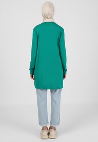 Tunique verte à manches longues en tissu lisse, dotée d'une coupe droite et d'un ourlet asymétrique, associée à un jean bleu clair retroussé et à des chaussures beiges.