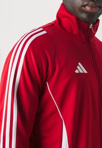 Rød atletisk jakke med hvide accenter, lynlås krave og tre hvide striber på skulderen. Har et lille gråt logo på brystet.