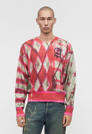 Mężczyzna w czerwono-beżowym swetrze w wzór argyle z gwiazdkami na rękawach i wyszytą literą "A" na piersi, w połączeniu z niebieskimi dżinsami.