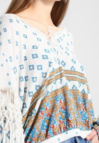 Biała boho bluzka z niebieskimi geometrycznymi wzorami, zdobiona brązowymi i pomarańczowymi akcentami, z dekoltem na sznurki i frędzlami na krawędziach.