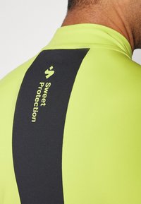 Kurtka w jaskrawozielonym kolorze z czarnym paskiem z logo "Sweet Protection". Gładka tekstura, idealna na aktywności na świeżym powietrzu.