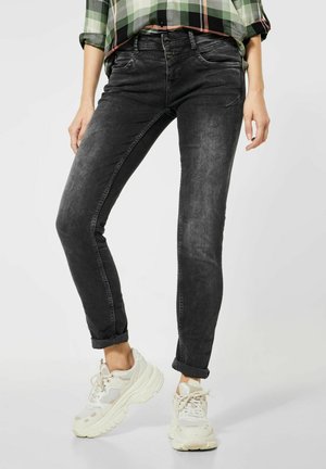 Vaqueros de denim negro con un corte ajustado, textura desgastada, con cierre de botón y cremallera, y puños enrollados, combinados con zapatillas blancas.