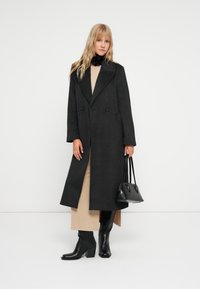 Pitkä musta villakangastakki, jossa on kaksirivinen malli, leveät kaulukset, päällä beige mekko, yhdistettynä mustiin korkokenkiin ja pieneen käsilaukkuun.