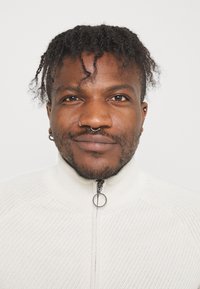 Bílá zipová mikina s texturovaným vzorem, s vysokým límcem a kruhovým stahovákem na zip. Záber z blízka na osobu s krátkými dredy.