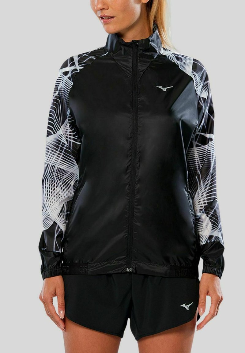 Schwarze leichte Jacke mit weißen abstrakten Mustern an den Ärmeln, Reißverschluss vorne und elastischen Bündchen. Kombiniert mit schwarzen Shorts.