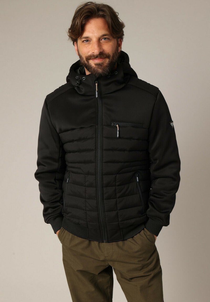 Homme avec une barbe portant une veste matelassée noire à capuche et un pantalon vert olive, se tenant les mains dans les poches contre un fond uni.