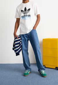 Bílý bavlněný tričko s černým logem "adidas" a modrými akcenty, spárované s modrými džíny a zelenými teniskami s bílými pruhy.