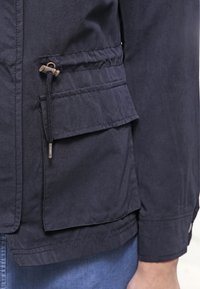 Granatowa kurtka wykonana z gładkiej tkaniny, wyposażona w regulowany sznurek w talii oraz klapową kieszeń po lewej stronie z metalowymi ozdobami.