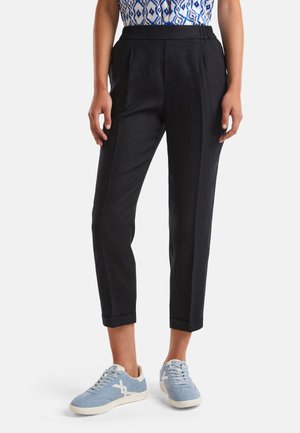 Kobieta w czarnych krótkich spodniach, niebiesko-białej bluzce z wzorem oraz jasnoniebieskich trampkach, stojąca na białym tle.