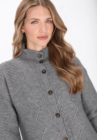 Szary dziany kardigan z wysokim kołnierzem, z fakturowanymi wzorami w zygzak, dużymi ciemnymi guzikami i ściągaczowymi rękawami.