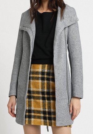 Femme portant un manteau gris zippé par-dessus un top noir et une jupe à carreaux jaune et noire, debout devant un fond uni.