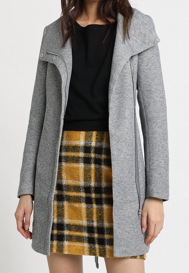 Femme portant un manteau gris zippé par-dessus un top noir et une jupe à carreaux jaune et noire, debout devant un fond uni.