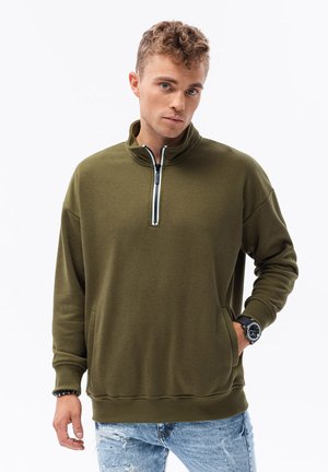 Sudadera con cremallera de color verde oliva, con cuello alto, puños y dobladillo de canalé. Cuenta con un bolsillo frontal y una tela texturizada. Se lleva con jeans azules desgastados.