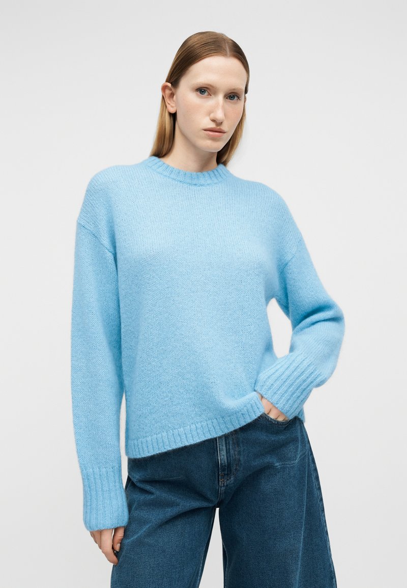 Frau mit glattem Haar, die einen hellblauen Strickpullover und blaue Jeans trägt, steht mit einer Hand in der Tasche vor einem einfarbigen Hintergrund.