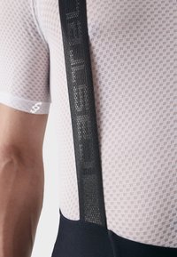 Maillot de cyclisme en maille blanche avec un motif texturé, featuring des bretelles ajustables noires et une ceinture en tissu sombre.
