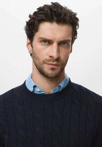 Jersey de punto en azul marino sobre una camisa de cuello azul claro; presenta un cuello acanalado y un patrón texturizado para un detalle adicional.
