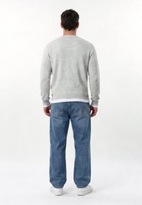 Άντρας που φοράει ανοιχτό γκρι πουλόβερ, μπλε τζιν και λευκά αθλητικά παπούτσια, στέκεται με την πλάτη προς τα εμάς σε απλό λευκό φόντο.