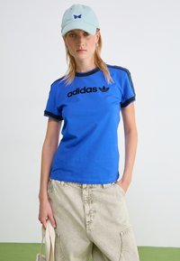 Blå bomulls t-skjorte med svarte detaljer og "adidas"-logo; kombinert med lyse, teksturerte baggy bukser og en mintgrønn caps.