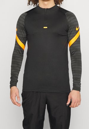 Homme portant un haut de sport noir à manches longues avec des manches grises à motifs et des bandes orange, associé à un pantalon de sport noir.