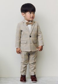 Light brown plaid suit with a single button jacket, matching trousers, white shirt, and a beige bow tie. Brown dress shoes complete the look.