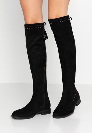 Black suede over-the-knee boots with side zipper, small silver studs around top and heel, worn by a standing person against a white background.