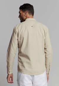 Beige langwerpige blouse met een eenvoudig design, een kraag met knopen en zichtbare knoopaccenten op de mouwen, gemaakt van ademende stof.