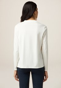 Camiseta blanca de manga larga con cuello redondo, textura suave y dobladillo recto, combinada con pantalones azul oscuro. Diseño sencillo, sin estampados.