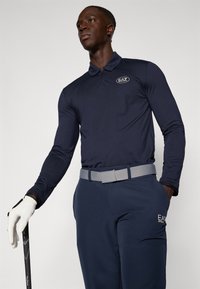 Mornarska polo majica z dolgimi rokavi iz teksturirane tkanine, ovratnikom in logotipom EA7. Ujemajoče se mornarske hlače in siva pas, ki drži golf palico.
