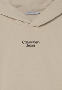 Beżowa bawełniana bluza z kapturem, z kieszenią kangurzą, kapturem ze sznurkiem i haftowanym logo "Calvin Klein Jeans" w czerni na piersi.