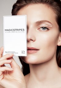 Magicstripes ooglidlifttape verpakt in een heldere rechthoek. Bevat tekst voor maten S, M en L. Een model houdt het pakketje dicht bij haar gezicht.