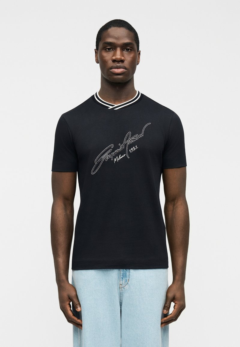 Άνδρας που φοράει μαύρο κοντομάνικο πουκάμισο μ ε λευκά γράμματα «Giorgio Armani Milano 1981» και ανοιχτό μπλε τζιν, στέκεται μπροστά σε απλό λευκό φόντο.