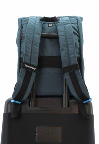 Mochila de tela azul con correas acolchadas unida a una maleta negra con ruedas. Presenta un parche con el logo y un asa de transporte, con un cochecito retráctil.