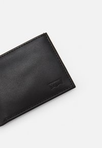 Portefeuille en cuir noir avec une texture lisse, des bords cousus et un petit logo en relief sur un côté. Forme rectangulaire.