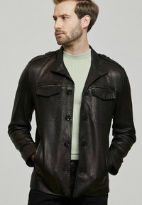 Zwarte leren jas met een licht gestructureerd oppervlak, knoopsluiting, twee borstzakken en een moderne snit. Gedragen over een lichtgroene trui.