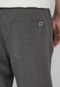 Grijze gemêleerde broek met een achterzak en een klein logo-label. Het materiaal lijkt geweven met een subtiel patroon.