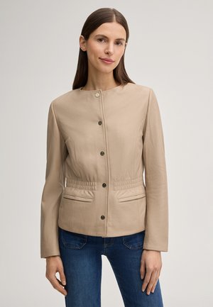 Kobieta ubrana w beżową kurtkę zapinaną na guziki z marszczonym stanem oraz niebieskie jeansy, stojąca na tle jednolitego jasnego tła.