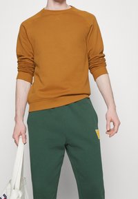 Sudadera de algodón naranja con puños y dobladillo acanalados, combinada con pantalones de chándal de algodón verdes que tienen un parche amarillo, sostenida con una bolsa de tela blanca.