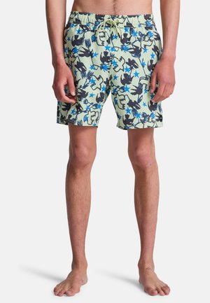 Mężczyzna w jasnych zielonych szortach kąpielowych z niebieskimi gwiazdami i czarnymi abstrakcyjnymi wzorami zwierzęcymi, stojący boso na jednolitym tle.