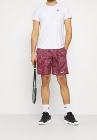 Vit kortärmad Nike-tröja med rutmönster, vinröda texturerade shorts, svarta skor och tennisrack. Märkbart logotyp på tröjan och shortsen.