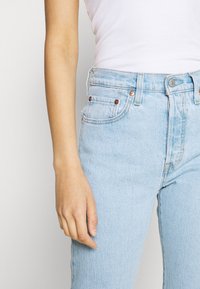 Jeans de mezclilla azul claro con cintura alta, que presentan remaches de cobre, bolsillos frontales y una textura ligeramente desgastada. Mano visible descansando en el costado.