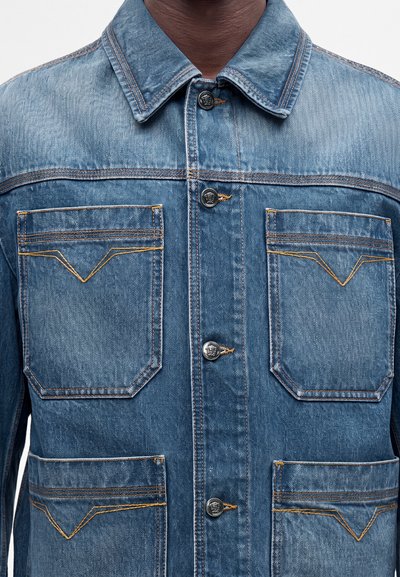 Gros plan d'une personne portant une veste en denim bleue avec quatre poches avant et des boutons métalliques au centre.