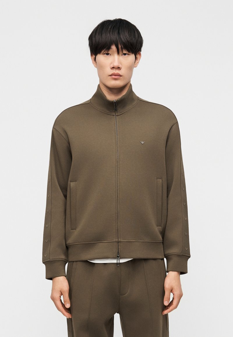 Άνδρας που φοράει χακί ζακέτα με φερμουάρ και παντελόνι στο ίδιο χρώμα, στέκεται μπροστά σε ένα απλό ανοιχτό φόντο με τα χέρια χαλαρά.