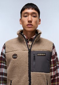 Beige fleece vest with gray front panel, zip closure, and logo patch. Worn over a plaid shirt with red, green, and beige colors.