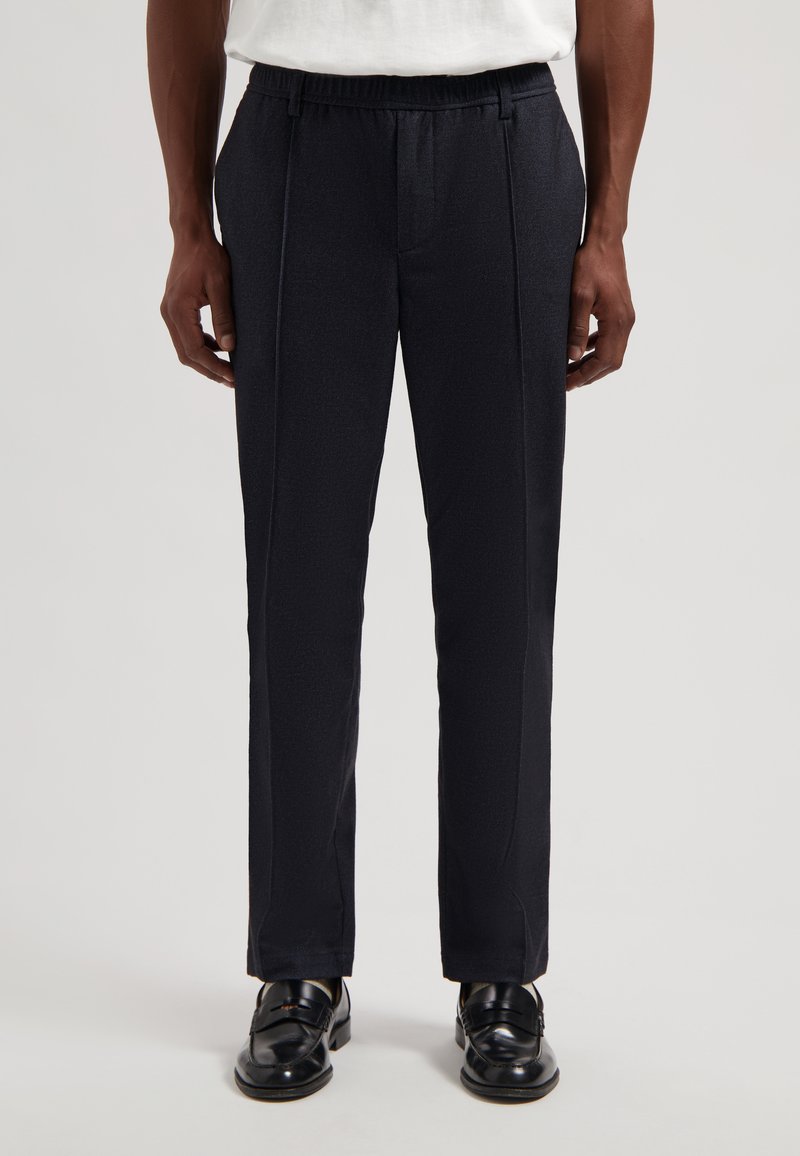 Granatowe spodnie na miarę z elastycznym paskiem w talii, o gładkiej fakturze i plisach z przodu, zestawione z czarnymi skórzanymi mokasynami.