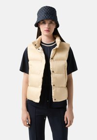 Beige gesteppte Puffer-Weste mit Druckknöpfen, getragen über einem navyfarbenen Oberteil mit gestreiftem Kragen. Accessoires sind ein dunkelblauer Bucket Hat.