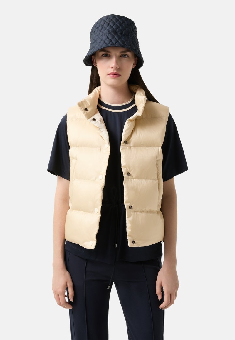 Beige gesteppte Puffer-Weste mit Druckknöpfen, getragen über einem navyfarbenen Oberteil mit gestreiftem Kragen. Accessoires sind ein dunkelblauer Bucket Hat.